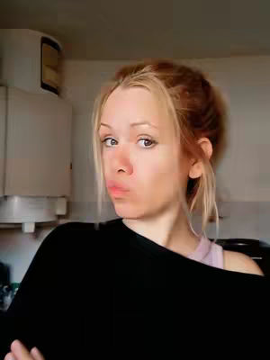 A selfie of a young woman with blonde hair pulled back. She's looking at the camera with a slight pout, wearing a black top and a light-colored tank top underneath. The background appears to be a kitchen setting with a water heater visible.