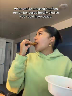A young woman with dark hair and a light green hoodie, bites into a cookie while holding a white bowl. Behind her is a light-colored ceiling with recessed lighting and part of a doorway visible. The lighting is warm, suggesting an indoor setting.