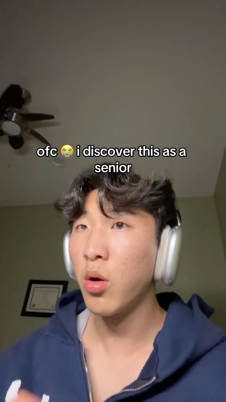 A young man with dark hair wearing a navy blue hoodie and white headphones is centered in the frame. He has a surprised expression, with slightly opened mouth and eyes looking slightly upward. Behind him, there's a blurry domestic setting visible: a ceiling fan, part of a wall, and a framed picture hanging on the wall. Text overlayed on the image says "ofc 😭 i discover this as a senior".