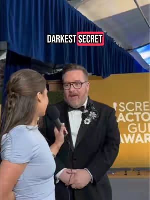 A woman with brown hair interviews a man in a tuxedo with a bow tie in front of a backdrop that says "Screen Actors Guild Awards". A text overlay reads "DARKEST SECRET".