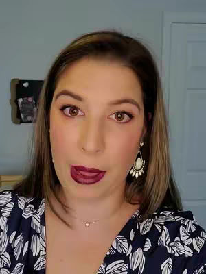 A woman with brown hair, dark lipstick, and a patterned shirt looks directly at the camera with her tongue out. The background includes a gray wall and a door.