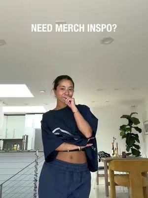 A woman stands indoors, posed in front of a bright room interior with a high ceiling. The woman is wearing a dark blue top and matching pants. The text: "NEED MERCH INSO?" is overlaid on the image.