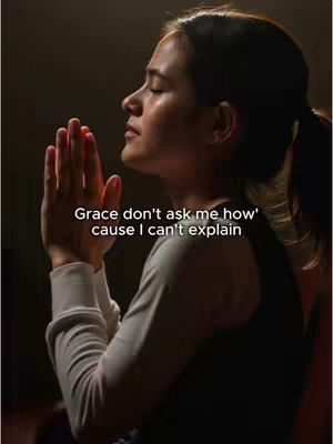 A woman with brown hair is depicted in a dimly lit setting, her hands clasped together in prayer, eyes closed, and head tilted slightly upward. Overlayed on the image is white text that reads "Grace don't ask me how' cause I can't explain". The overall composition suggests a moment of reflection, faith, and personal connection with spiritual undertones.
