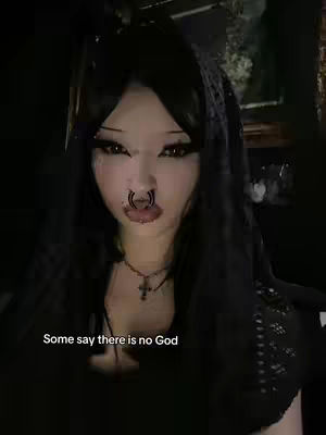 A young woman with dark hair and makeup is the central focus. She wears a dark veil and a nose ring. The image is dark, with dim lighting, and text at the bottom reads 'Some say there is no God'.