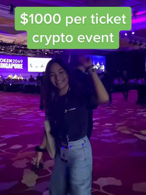 A smiling young woman stands in a large, ornately decorated room with reddish-brown patterned flooring and purple uplighting. Above her head, a green rectangular text overlay reads '$1000 per ticket crypto event'. The background includes a stage with a blue and purple backdrop, partially obscuring other people.