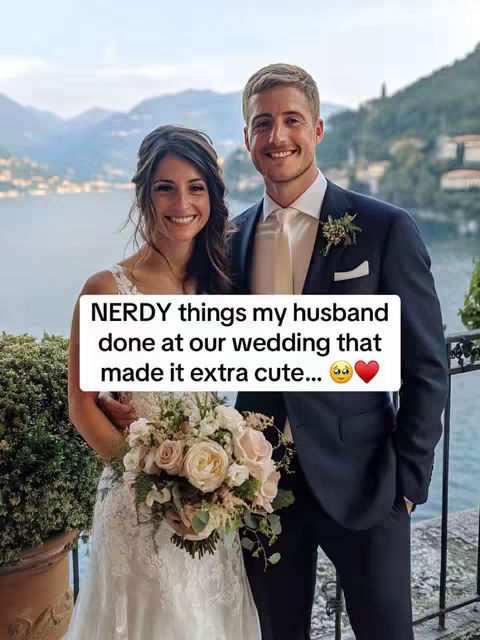 A smiling couple poses for a wedding photo outdoors. The woman on the left wears a white wedding dress and holds a bouquet. The man on the right wears a dark suit with a white shirt and tie. Behind them are a lake, mountains, and a slightly overcast sky.