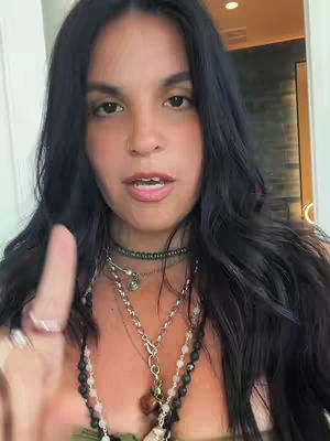 A woman with long, dark hair is shown in a close-up shot, appearing to be in a room with a light source visible. She is looking directly at the camera, with her mouth slightly open as if she is speaking. She is wearing several necklaces, and her hand is raised with the index finger extended. A ring can be seen on her finger. The overall composition highlights her face and upper body.