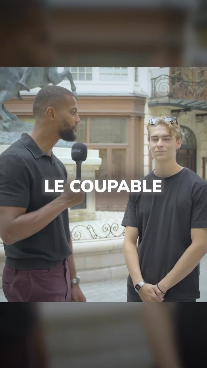 A wide shot shows two men in an outdoor setting. The man on the left is holding a microphone and facing the camera, while the man on the right is looking towards the interviewer or off-camera. The background features a building with architectural details and possibly a fountain.