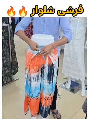 A person is shown adjusting a pair of brightly colored, tie-dye patterned pants. The pants are primarily in shades of orange, blue, and black, with a white central band. The person is wearing a light blue shirt. The background includes racks of clothing and mannequins.