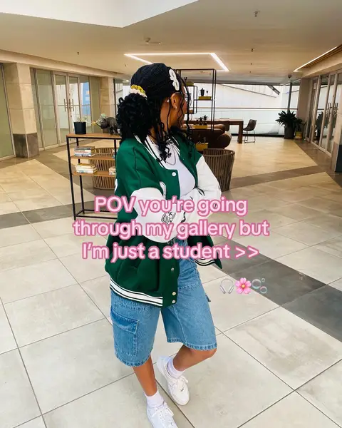 A young woman, viewed from behind and to the side, stands in a modern, open space with light-colored floors and walls. She is wearing a green and white letterman jacket, denim shorts, and white low-top sneakers. Her dark hair is styled with gel and a bun, and she wears glasses. The background shows a corridor with glass doors, a small table with items, and neutral-toned decor.