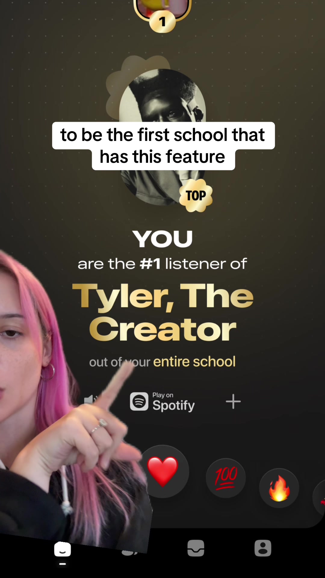 The cover image features a woman with pink hair on the left side, pointing at the center of the screen. The upper portion of the screen displays text overlay with text: "to be the first school that has this feature". Below that is a graphic stating "YOU are the #1 listener of Tyler, The Creator out of your entire school". The Spotify logo with a play button is visible at the bottom, along with interactive UI elements of a heart, a "100" emoji and a fire emoji. The background is a dark gradient with small light dots.