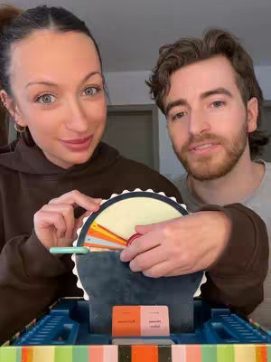 The cover image features two people, a woman on the left and a man on the right, seemingly interacting with a game or device in front of them. The woman is looking towards the camera with a slight smile, and the man has a neutral expression. They are both holding parts of a circular game mechanism featuring colors and designs. The background is blurred a bit.