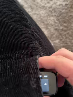 The image shows a close-up view of a black corduroy fabric. A person's hand is visible, reaching into a pocket. In the pocket, a smart device with a black screen displaying an interface with a few icons is partially visible. The background is a soft, blurred texture, likely a carpet or a similar fabric.
