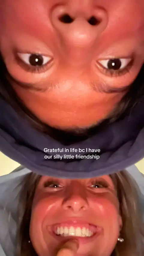 Two women's faces are close together, each taking up a large portion of the frame. The top face is angled downwards, showing the nose and eyes with the rest obscured by a blue hoodie. The bottom face is smiling widely with a finger touching the lips and wearing a blue hoodie. Text is overlaid in the middle of the image.