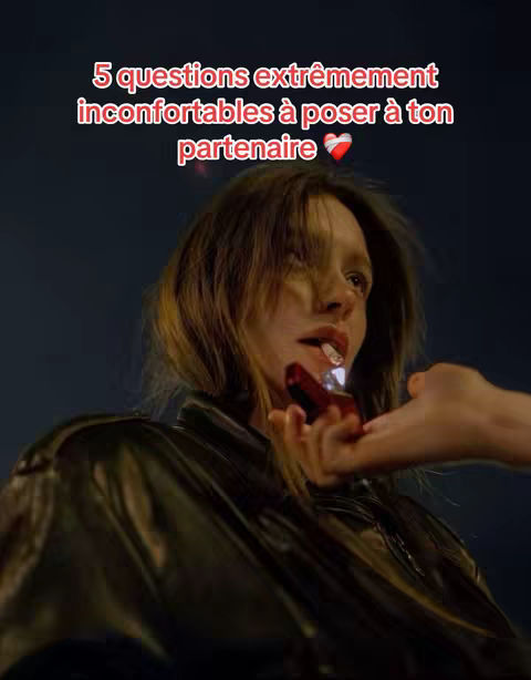 A medium shot shows a woman gazing off-camera with a lit object in her mouth, possibly a cigarette or something similar. Her hair is shoulder-length and tousled. A hand is extending towards her, holding an object that appears to be a small, rectangular case or lighter. The background is a dark, blurred gradient.