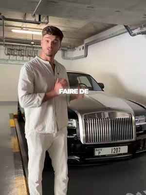 A man wearing a white shirt and pants stands leaning forward in a parking garage with a luxury car behind him. The words "FAIRE DE" appear above the car.