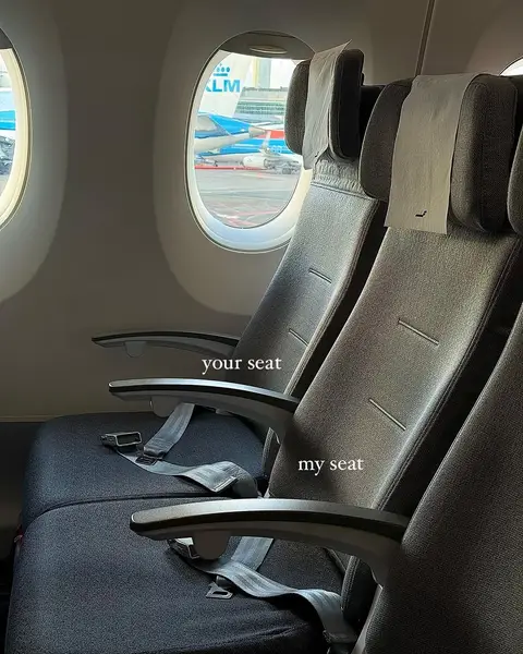 An interior shot of an airplane cabin with three seats visible. The seats are gray with headrests and seatbelts. The windows show an airport scene outside with a KLM airplane visible.