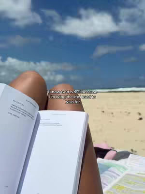 A woman lies on a beach reading a book. The open book is in the foreground, and the text overlay appears above it. The background shows the ocean and blue sky.