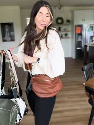 A woman with long brown hair, styled with blonde highlights, smiles broadly while looking slightly to the right. She wears a cream-colored, ruffled button-up shirt and black pants, and is holding multiple bags. The bags hang down in front of her. Behind her, the background shows a bright living area, with a doorframe, and a glimpse of a kitchen.
