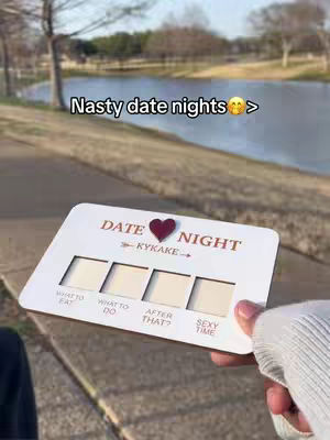 A hand holds a white card outdoors against a backdrop of a lake, trees, and a grassy area. The card features pre-printed text and spaces for customization. The sky is overcast, creating diffused natural lighting.