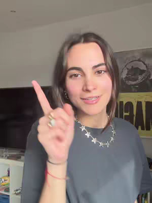 A woman with long brown hair and braces smiles at the camera, pointing her index finger upwards. She wears a dark gray t-shirt and a silver star necklace. Jewelry includes a few rings and various bracelets, and her facial expression is friendly.