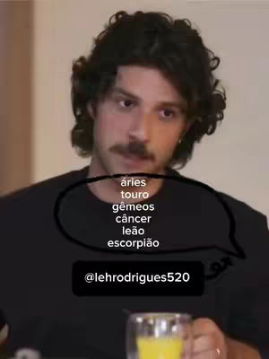 A man with dark curly hair and a mustache is shown, holding a clear glass mug with a yellow liquid. Behind him, a neutral wall serves as the background. Overlaid on the image is a speech bubble containing a list of astrological signs in Portuguese, and below that is a black rectangular box with a username.