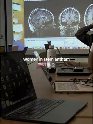 The image shows a dimly lit room with a laptop in the foreground. A projection screen in the background displays several MRI brain scans. In front of the screen sits a white water bottle and other supplies. The text overlay says 'women in stem anthem'.