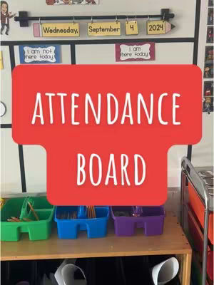 The image showcases a classroom attendance board. A whiteboard has a horizontal bar with hanging labels indicating the day, date, and year. Below, a large red rectangle with 'ATTENDANCE BOARD' in white text dominates the center. The lower portion of the image reveals shelves with colorful bins, suggesting an organized and functional learning environment.