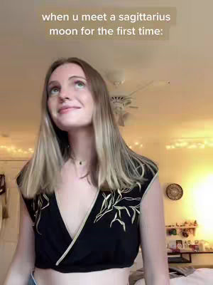 A young woman with blonde hair looks up with a slight smile in a well-lit room. The background includes a ceiling fan and soft lighting, suggesting a casual indoor setting. The woman wears a dark top with leaf-like patterns. Text overlayed at the top says, 'when u meet a sagittarius moon for the first time:'