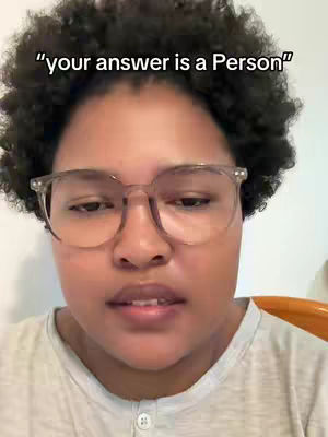 A selfie-style shot featuring a person with dark, curly hair, wearing clear-framed glasses. The person is centered in the frame, with a neutral expression and relaxed pose. The lighting appears soft, and the background is a plain white wall with a glimpse of an orange surface on the right.