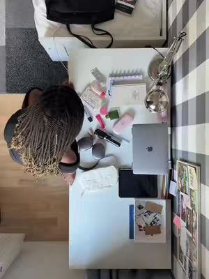 The overhead shot shows a desk setup with various items, including a laptop, headphones, cosmetics, and personal items like cards and what appears to be a microphone. A person with braided hair is framed at the lower left corner of the image, partially visible. The desk is white, and the background includes a patterned wall and part of a bed.