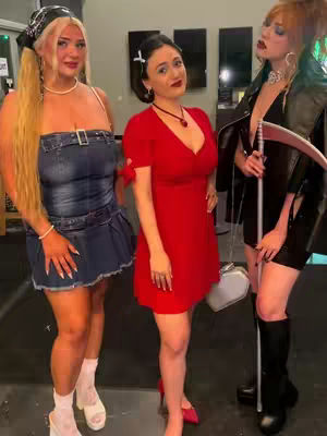 Three young women in various costumes are posing side by side in a well-lit environment, likely indoors. The woman on the left wears a denim dress and has blonde hair styled in two high ponytails. The middle woman is dressed in a red dress. The woman on the right wears a black dress, a light jacket, and holds a scythe. The background features dark panels and decorative elements
