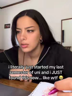 A young woman with long, dark hair is looking towards her left with a concerned expression. She wears a black zip-up top. Her right hand is scribbling on a notepad in front of her. The background is a bright room with a painting and a mirror.
