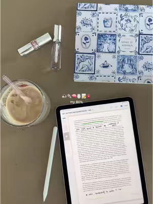 An overhead shot of a table with a light brown surface. On it, there's a clear glass with a brown liquid and a straw, next to a tablet displaying text with a stylus beside it. Further up, a patterned fabric is visible, below some beauty products, offering a sense of a workspace.
