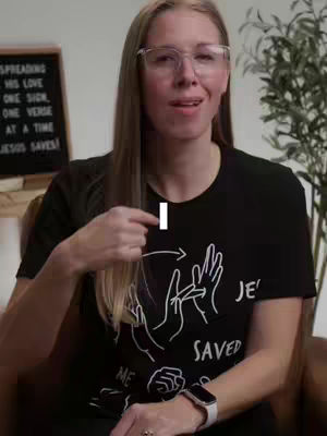 A medium shot shows a woman with long blonde hair, wearing glasses and a black t-shirt, gesturing with her right hand. Behind her, a chalkboard displays a religious message. To her right, a blurred plant adds depth. The image is well-lit and conveys a message-oriented atmosphere.