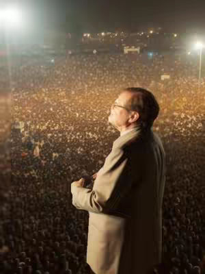 The image shows a man, likely a preacher, standing and looking upwards. A large crowd, possibly a congregation, fills almost the entire background. At the top left, there is a strong light source, perhaps a spotlight. The overall tone of the image seems to be one of reverence and faith.