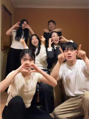 A group of seven people, primarily young adults, are gathered closely together, posing for a photo with a warm, inviting ambiance. The composition features a blend of formal and casual attire. Some individuals are seated in the foreground, while others are positioned toward the back. The overall color scheme is natural, incorporating soft tones. The lighting appears to be ambient, producing a warm, indoor setting. The pose of the group shows a moment of affection and playfulness, conveying a sense of joy and unity.