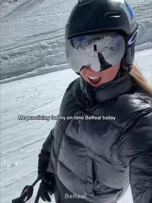 A selfie shot shows a person wearing a black ski jacket and helmet with mirrored goggles against a snowy mountain background. The person is mid-action, mouth open, suggesting they are speaking or reacting. The angle is from slightly below, looking up at the person.