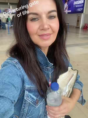 A woman with dark hair smiles at the camera in what appears to be an airport. She wears a denim jacket and holds a water bottle and a notepad. The setting suggests travel or a moment of departure.