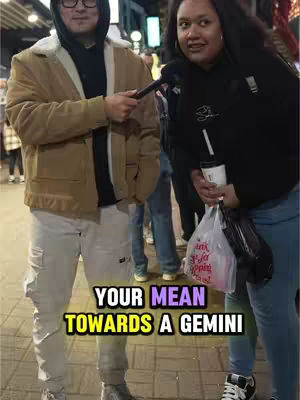 Two people are facing the camera on a city street, discussing astrology. The person on the left, wearing a coat and a hoodie, is holding a microphone, and the person on the right is holding a drink. Text overlayed on the bottom explains the topic.