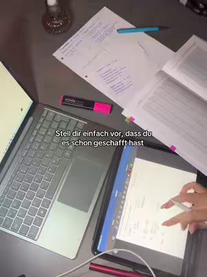 Overhead shot of a desk with a laptop, tablet, books, and study materials. A hand is visible, using a stylus on the tablet. The overall scene suggests study or work.