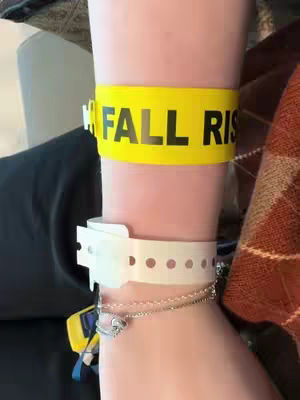 The image shows a close-up of a person's wrist and forearm. A bright yellow band with the words "FALL RISK" in large black letters encircles the arm. Below this, a white hospital wristband is visible, and a silver bracelet with a charm hangs from the wrist. The skin tone is light, and the overall image suggests a medical or hospital setting.