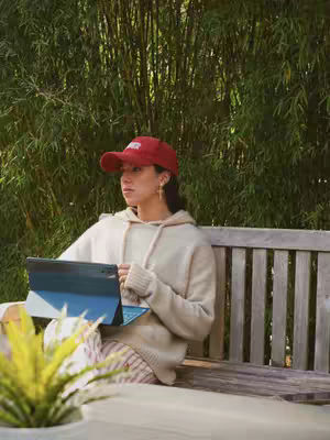 A woman wearing a red cap and hoodie sits on a bench, holding a tablet. She is in a garden setting, with a lush, green backdrop. The image has a natural, well-lit appearance.