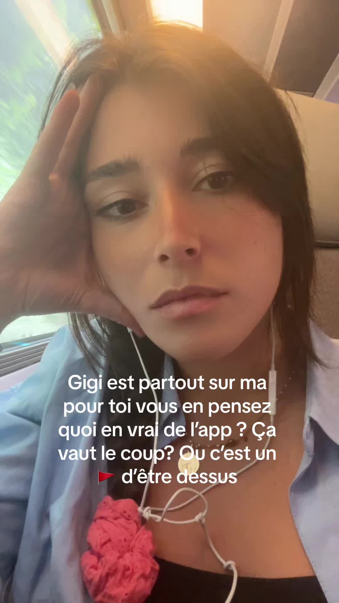 A selfie showing a young woman with dark hair, her face framed in the center. Her eyes are looking down, and her hand rests on her cheek. She wears a light blue top with a pink object at her neck. The background includes blurry elements suggesting an indoor setting with cool light. Text is overlaid on the bottom part of the image, written in French.
