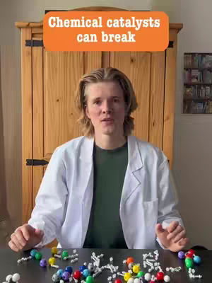 A man in a white lab coat stands facing the camera with a table in front of him covered in colorful model molecular structures. Above his head is an orange rectangle with white text.