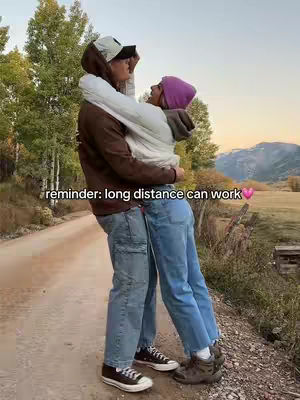 A couple is embracing on a dirt path with trees and mountains in the background. The man holds the woman in his arms. The text overlay reads, "reminder: long distance can work." The overall composition appears warm and romantic.