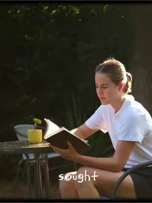 A young woman sits at a small table reading a book outdoors. The background features lush green foliage, suggesting a garden setting. The sunlight creates highlights on her hair and the book, and adds to the image's peaceful feel.