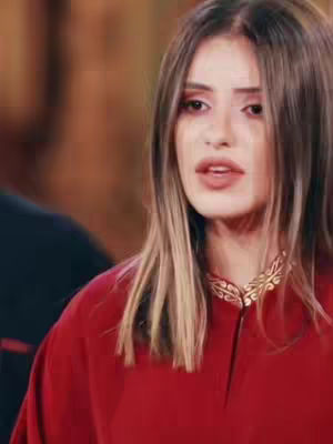 A medium shot features a woman with light brown hair and fair skin, looking slightly to the side with a soft expression. She is wearing a red top with a gold neckline. The background is blurred, featuring the suggestion of an interior.