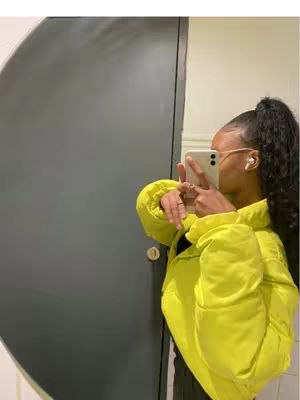 A person takes a selfie, mirroring in a cabinet with a mirror and wearing a neon yellow jacket with the phone in their hand. They have dark hair in a high ponytail, and white earbuds in. The background includes the gray cabinet and the light wall.