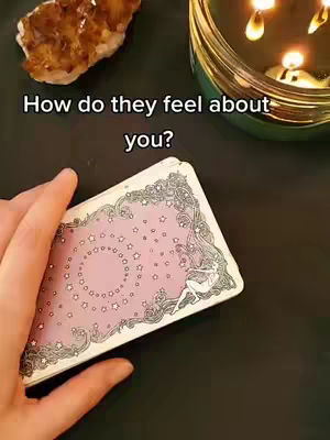 The image shows a tarot card reading setup. A hand holds a deck of tarot cards. On the background, a lit candle sits in a decorative glass container and a stone. The cards are pink and white with star-themed designs on the back, and the text overlay suggests a question about feelings.